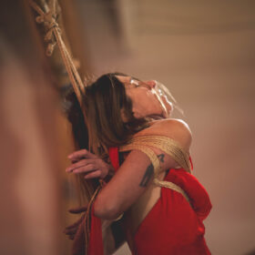 Nawataneko kinbaku model and performer being tied in Gote hands on the back in the red dress presenting in San Francisco