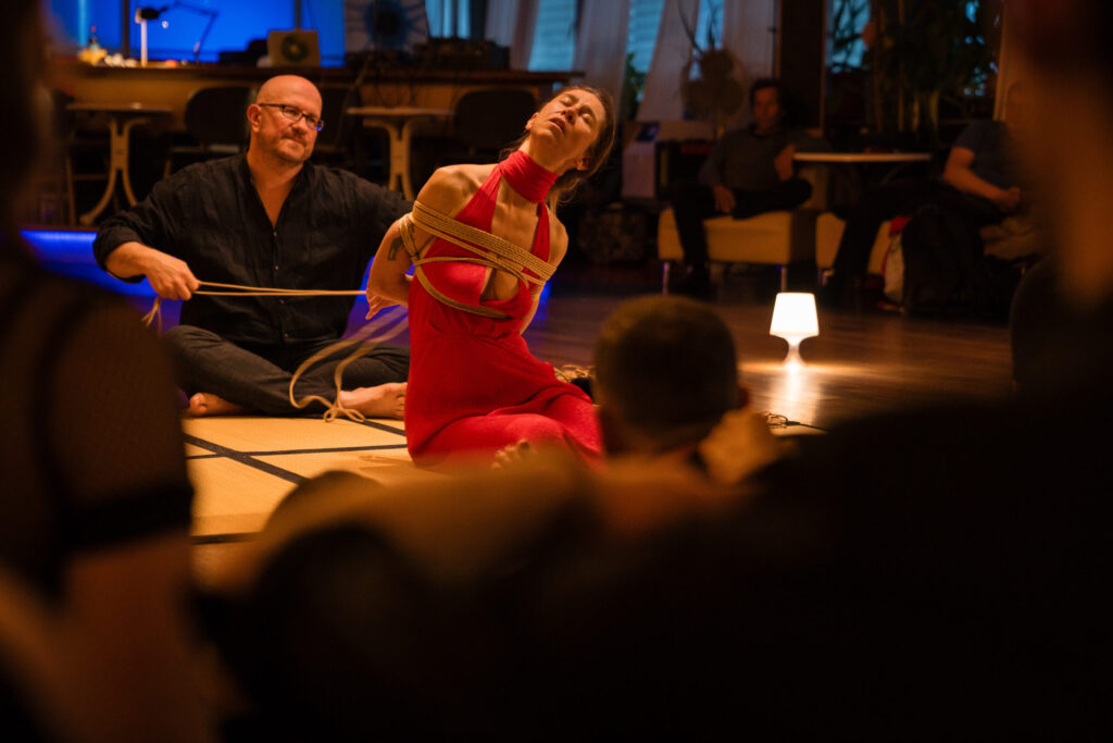 Rigger tying a rope bottom in a red dress during a Kinbaku performance in front of an audience